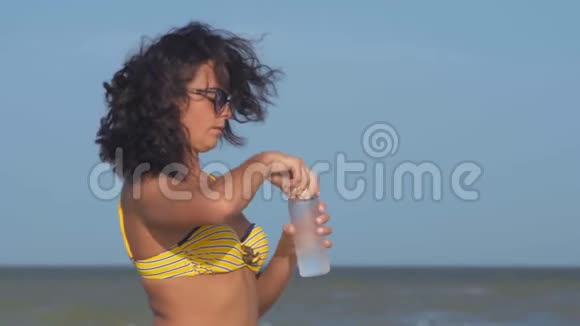 阳光眼镜中的贴身女人从海滩上的玻璃瓶中喝水视频的预览图