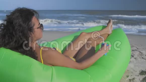 太阳眼镜中的女人躺在海滩上手里拿着鸡尾酒看着大海视频的预览图