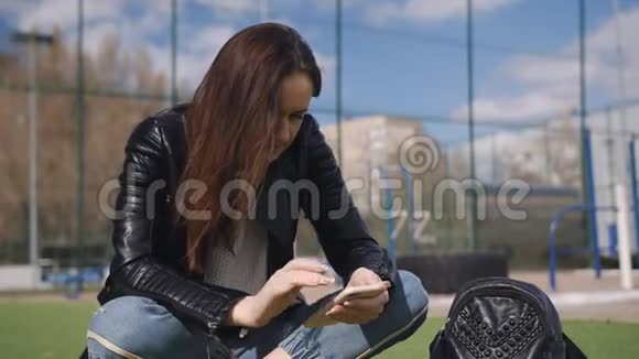女人穿着黑色皮夹克坐在操场上的草坪上用智能手机视频的预览图