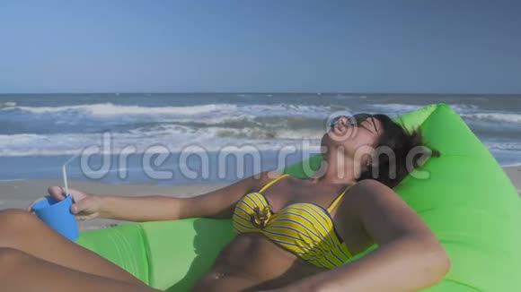 近景美丽的女人在比基尼日光浴海滩谎言绿色充气枕头与鸡尾酒在手视频的预览图