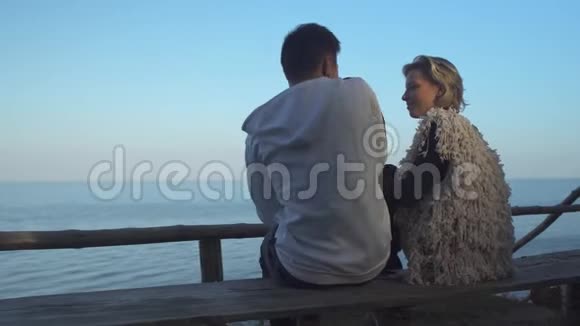年轻人和女孩坐在长凳上拥抱在水面的背景上年轻一对相爱的情侣视频的预览图