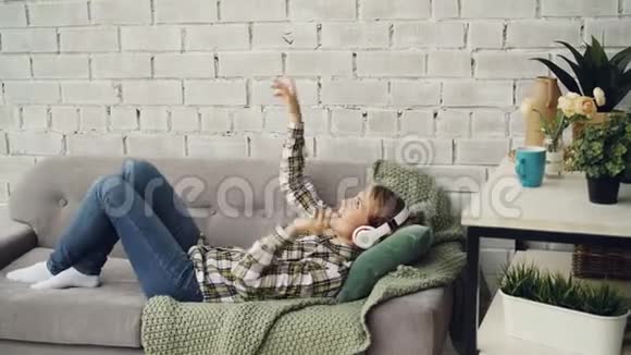 开朗的女孩正在听音乐戴着耳机移动着手臂和身体躺在家里的沙发上现代视频的预览图