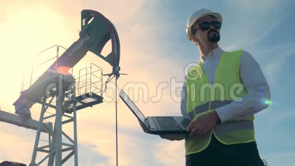 阳光普照的燃料井架和一名男性工程师在其前面操作一台笔记本电脑石油工业石油工业石油部门视频的预览图