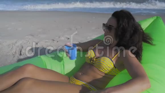在海滩上的绿色充气枕头上躺着阳光眼镜中的美丽女人并喝一杯鸡尾酒视频的预览图