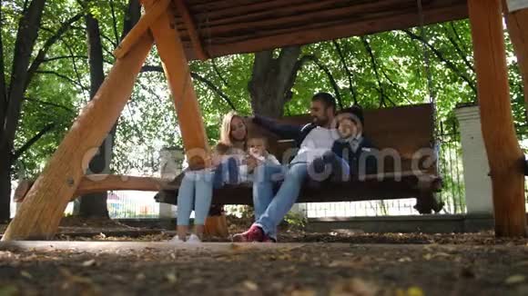 幸福的年轻家庭父亲母亲儿子和小女儿骑着秋千在公园里笑着笑4k慢慢视频的预览图