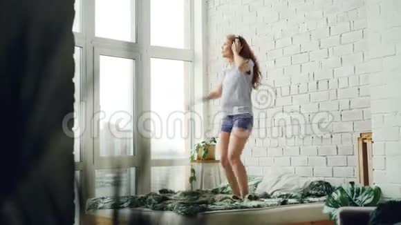 快乐的年轻女子戴着耳机在家里的床上跳舞享受着音乐双人床砖墙全景视频的预览图