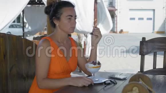 美丽的女人穿着橙色的裙子坐在海边的咖啡馆里吃着玻璃碗里的甜点视频的预览图