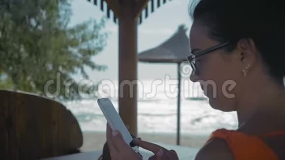 近景戴眼镜的美女坐在海边的咖啡馆里看着智能手机上的照片视频的预览图