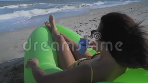 阳光下美丽的女人看着大海在绿色充气枕头上喝躺着的鸡尾酒视频的预览图
