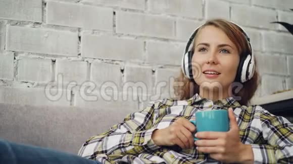 放松的女学生正在听音乐戴着耳机唱歌拿着一杯咖啡坐在阁楼的沙发上视频的预览图