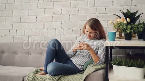 穿休闲服装的漂亮女孩使用智能手机坐在现代阁楼式公寓的沙发上面带微笑视频的预览图