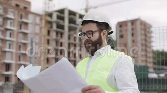 一位成功的年轻工程师或建筑师戴着白色头盔的肖像在他的建筑图纸中寻找视频的预览图