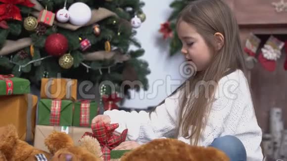 一个棕色长发的小女孩坐在圣诞树旁打开礼物视频的预览图