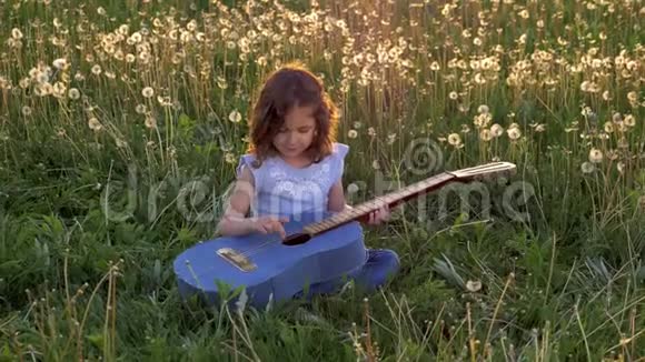 有趣迷人的小女孩坐在草地上弹牛仔裤吉他日落时盛开的蒲公英田野视频的预览图