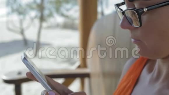 特写镜头戴眼镜的美女在坐在海边的咖啡馆里的智能手机上打印文字视频的预览图