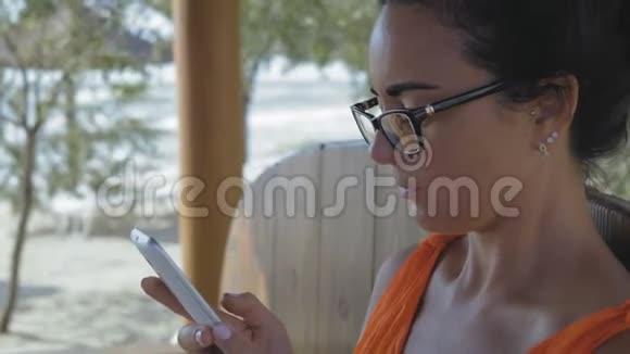 一位戴眼镜智能手机的美女坐在海边的咖啡馆里视频的预览图