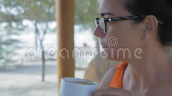 特写镜头戴眼镜的美女喝着咖啡坐在海边的咖啡馆里视频的预览图