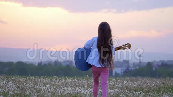 在城市风景的背景下滑稽的小女孩音乐家戴着太阳镜弹着吉他在丹德的草地上散步视频的预览图