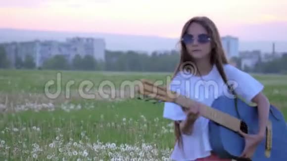 太阳眼镜上戴着嬉皮笑脸的小女孩在日落时在城市附近的户外弹吉他情感和模仿流行艺术家视频的预览图
