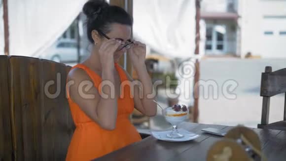 美丽的女人穿着橙色的裙子坐在海边的咖啡馆里摘下眼镜吃甜点视频的预览图