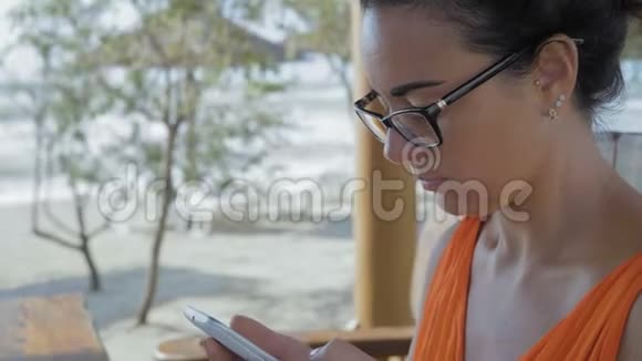 特写镜头戴眼镜的美女坐在海边的咖啡馆里在智能手机上写了一篇文章视频的预览图