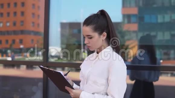 穿着白衬衫的年轻商务小姐站在商务中心附近看工作文件视频的预览图