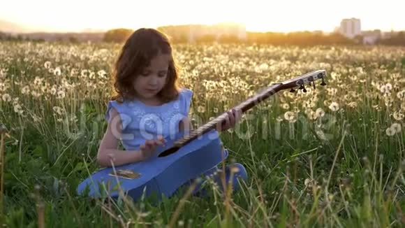 小女孩坐在草地上弹牛仔裤吉他在蒲公英的田野里春天在日落时绽放自然概念音乐视频的预览图