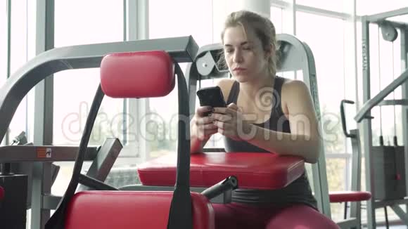 年轻女子坐在健身房用智能手机视频的预览图