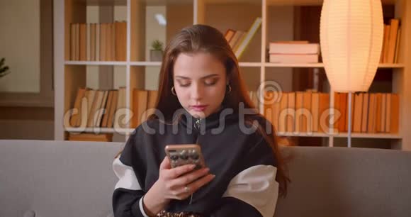 年轻少女坐在沙发上看手机的肖像家里有书架背景视频的预览图
