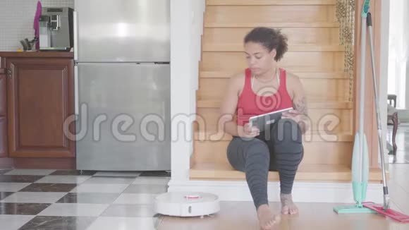 非洲裔美国妇女坐在大房子一楼的楼梯上在她的平板电脑上发短信现代机器人真空视频的预览图