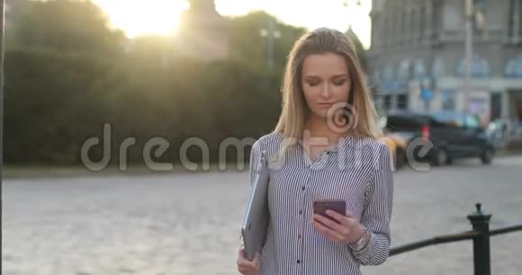 一个穿着衬衫的迷人金发女郎拿着笔记本在手机上发短信她摸了摸她的头发视频的预览图