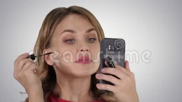 女人在睫毛上涂上黑色睫毛膏在渐变背景下看着她的手机视频的预览图