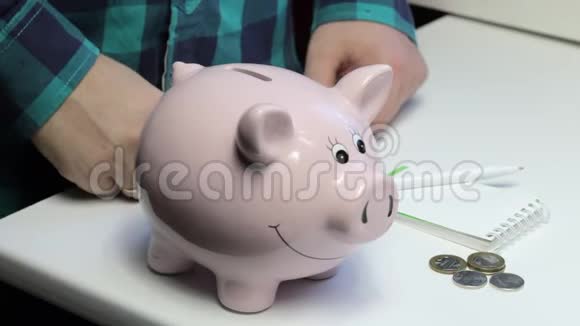 桌子上有一只粉红色的猪形的储蓄罐那个人手里拿着一枚硬币他把它依次放进一只小猪b里视频的预览图