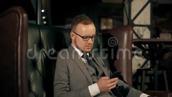 成功的胡子商人穿着西装坐在豪华的皮革沙发上看着电话4K段视频的预览图