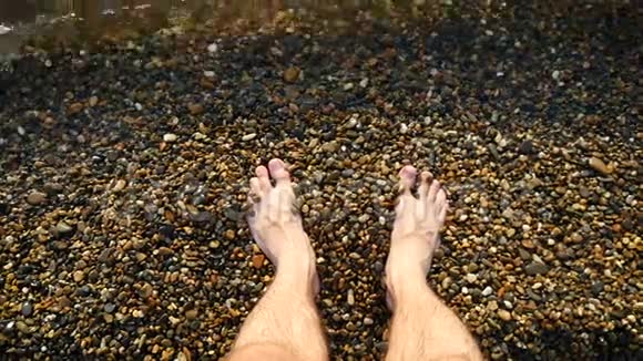 一个赤脚的人站在水中的鹅卵石海岸上照相机的焦点在那个人脚上视频的预览图