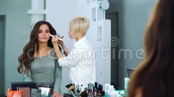 化妆师在美容院将粉涂在年轻女子脸上的镜面反射视频的预览图