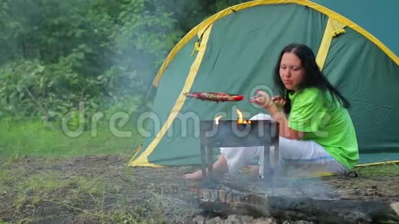 一个穿着绿色T恤的女人坐在森林里的帐篷里炉火正在她的胸罩里燃烧视频的预览图