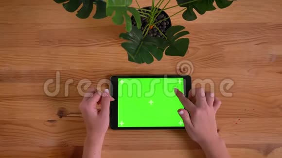 在木桌和植物背景上水平平板的绿色屏幕上打字的女子手的顶部镜头视频的预览图