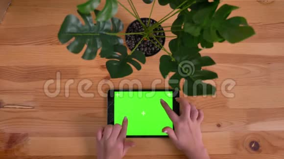 在木桌和植物背景上水平平板电脑的破绿色屏幕上打字的女人手的顶部镜头视频的预览图