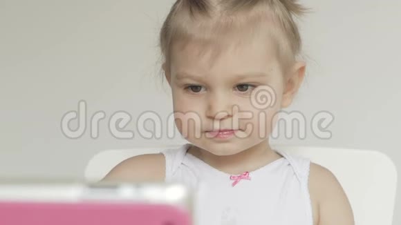 一个小女孩坐在孩子们桌子旁用勺子吃粥看着电子药片的屏幕视频的预览图