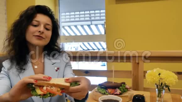 布鲁内特商务小姐穿着一件蓝色夹克在一家有黄色墙壁的舒适咖啡馆里吃个新鲜的茂盛的三明治视频的预览图