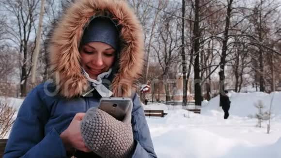 在冬天的公园里雪天里一位贴身的快乐女人坐在长凳上浏览着手机视频的预览图