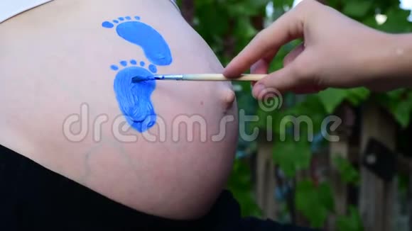 手用刷子画在腹部蓝色婴儿脚印上怀孕的概念特写视频的预览图