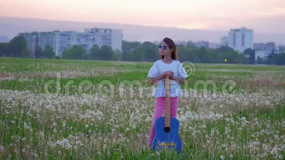 戴着太阳镜的年轻音乐家女孩站在球场上晚上在背景城市景观中拿着蓝色吉他灵感概念视频的预览图