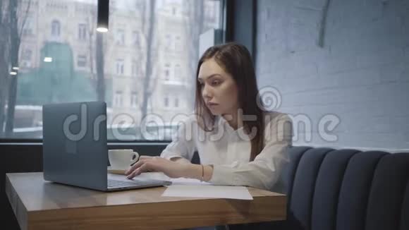 专心致志的女人用她的笔记本电脑坐在咖啡馆的桌子上女孩坐在靠近窗户的沙发上端着一杯视频的预览图