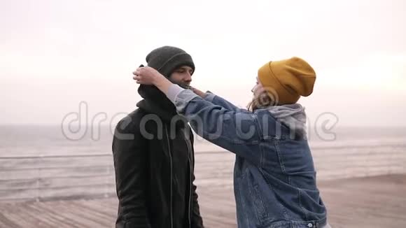 年轻可爱的一对夫妇穿着温暖的衣服和帽子一边站在海边一边一边有趣地纠正他们的帽子视频的预览图