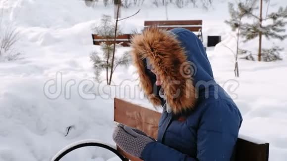 白天雪天在冬天的城市公园里女人坐在长椅上浏览手机视频的预览图