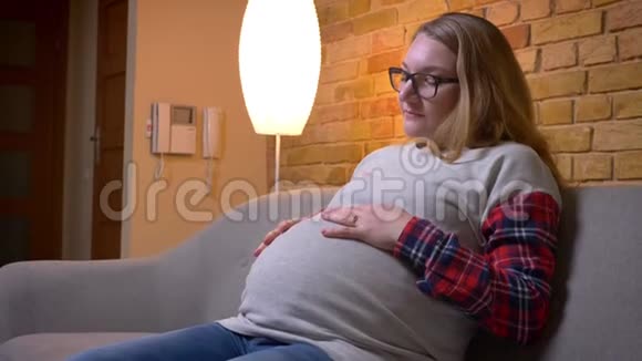 年轻怀孕女性在舒适的室内公寓里坐在沙发上轻轻抱着肚子的特写镜头视频的预览图