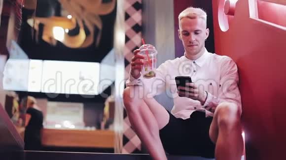 晚上使用智能手机的年轻魅力男子他坐在夜店里靠近霓虹灯招牌喝着鸡尾酒视频的预览图