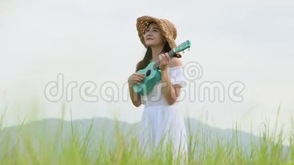 亚洲快乐的女人穿着白色连衣裙在绿色的草地上弹着四弦琴吉他夏天有着山山的背景音乐美人视频的预览图